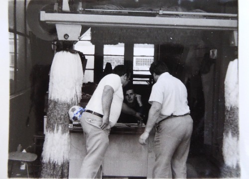 Auf dem schwarz-weiß Foto ist eine ehemalige Waschanlage, welche als Büro genutzt wird, zu sehen. Vor einem Tisch stehen zwei ältere Männer. Hinter dem Tisch sitzt Can.