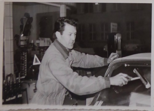 Auf dem schwarz-weiß Foto ist Can in einer Autowerkstatt zu sehen. Er wischt mit einem Scheibenwischer die Frontscheibe eines Autos.