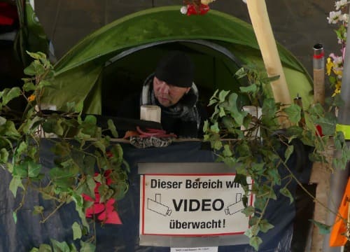 Auf dem Foto zu sehen ist Björn in seinem Zelt. Aus diesem schaut er raus. Im unteren Abschnitt des Bildes sieht man ein Schild mit der Inschrift: „Dieser Bereich wird Video überwacht.“. 
