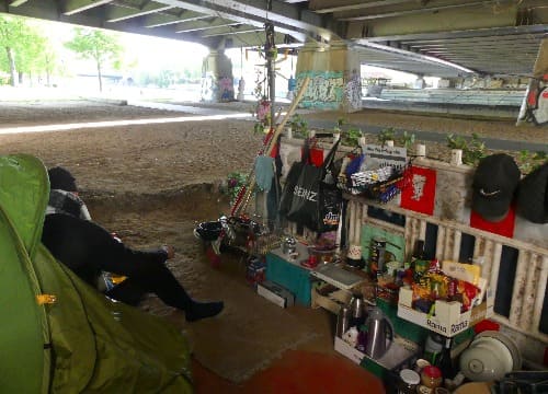 Auf dem Foto ist Björns selbst eingerichtete Ecke zu sehen. Diese ist mit verschiedenen Haushaltsartikeln wie Küchenutensilien und Lebensmitteln beladen. Links im Bild sitzt Björn vor seinem Zelt.